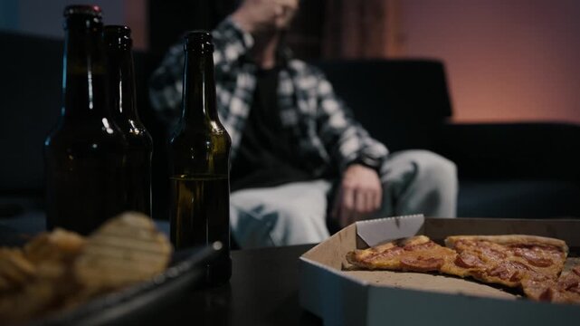 Man's hand grabs pizza slice from box, eats it and washes down with beer. Chips and bottles sharp in foreground, blurred man on couch behind. Moody game night at home.
