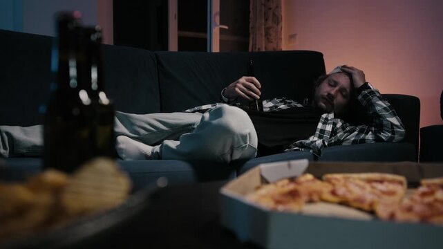 Man wakes up on couch with surprised reaction to TV, raises up and drinks beer. Pizza and chips blurred in foreground. Moody blue warm light. Lazy relatable solo night at home.
