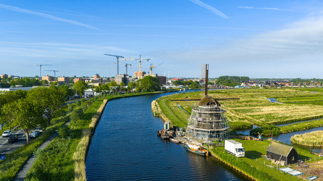Aerial view of the Oosterdel nature reserve featuring a historic windmill under renovation beside a winding canal and lush green polder fields Broek op Langedijk, Noord-Holland, Netherlands.