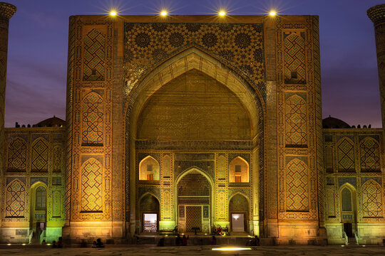 Illuminated Ulugh Beg Madrasah facade on Registan Square in Samarkand at twilight. Intricate tilework, a monumental iwan glow under warm lights against a deep blue sky. Ideal for travel publications.