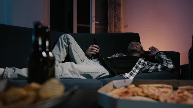 Young man asleep on couch still holding beer bottle, pizza and chips blurred in foreground. Moody TV glow. Relatable passed out after game night at home.
