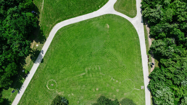Aerial view of a manicured green lawn with mowed patterns surrounded by lush forest trees and a winding white pedestrian path in Novi Sad, Vojvodina, Serbia.