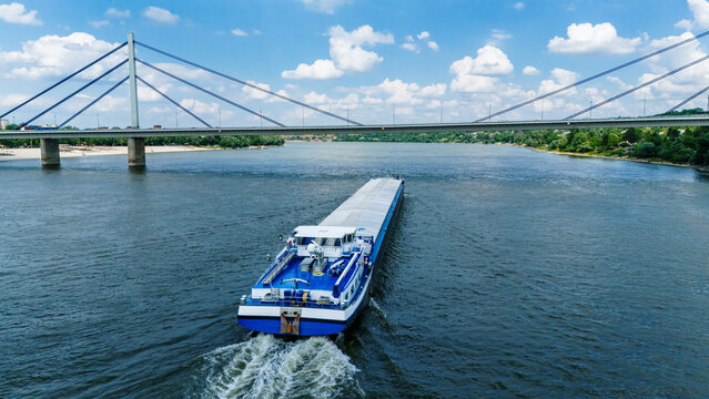 Aerial view of a large cargo barge navigating the Danube river towards the Liberty Bridge under a blue sky with white clouds in Novi Sad, Vojvodina, Serbia.