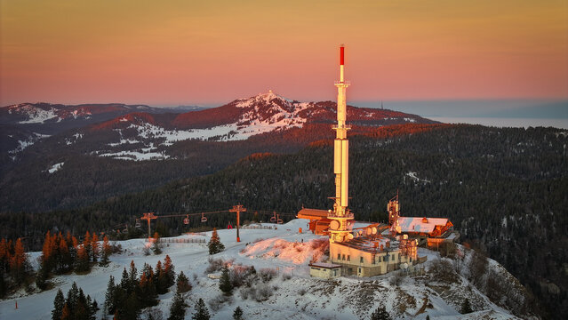 Aerial view of the Col de la Faucille telecommunications tower and ski lifts on a snowy mountain peak during a vibrant sunset in Gex, Auvergne-Rhone-Alpes, France.