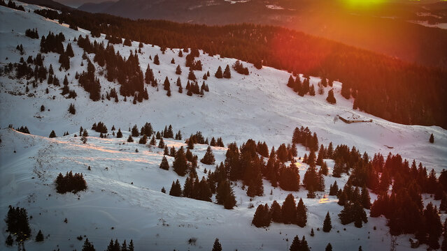 Aerial view of Col de la Faucille mountain pass covered in snow with pine trees and a small chalet during sunset in Gex, Auvergne-Rhone-Alpes, France.
