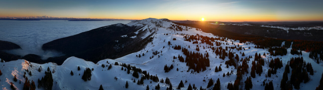 Aerial view of Col de la Faucille with snow-covered mountain ridges and pine trees above a sea of clouds at sunset Gex, Auvergne-Rhone-Alpes, France.