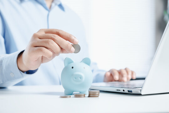 Business professional putting coin into piggy bank beside laptop with stacked coins on desk, personal savings and digital banking concept in soft blue corporate workspace with modern finance lifestyle