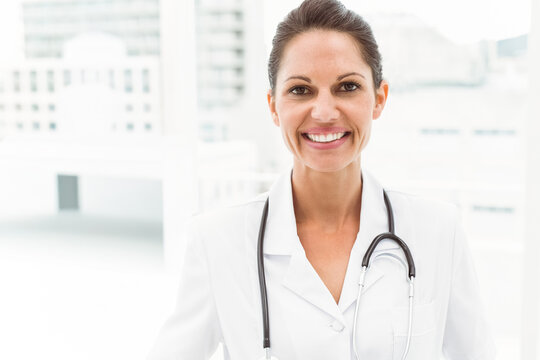 Mid adult female doctor standing smiling in clinic wearing labcoat stethoscope Vneck large windows