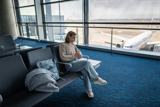 Female traveler sitting alone in airport departure lounge near panoramic windows while waiting for boarding looks at watch, afraid to miss flight. Travel moment, air transportation, solo journey trip.
