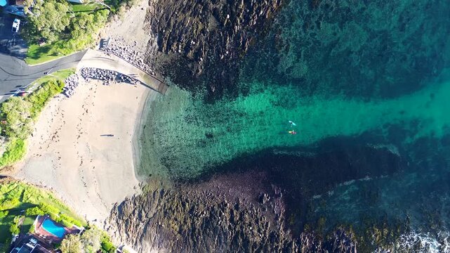 Drone aerial landscape of two kayakers waiting in a sandy beach cove surrounded by rocky reef coastline and public boat ramp at Lilli Pilli town suburbs South Coast NSW Australia tourism and leisure
