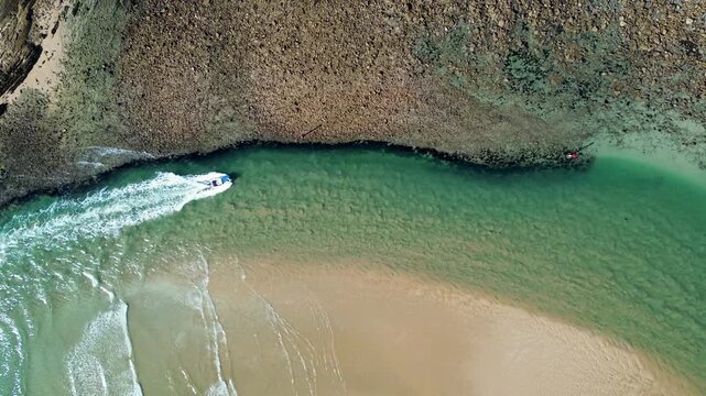 Drone aerial landscape of a speedboat travelling along the Tomaga River flowing channel inlet beside rocky headland terrain and sandy Tomakin Beach at Mossy Point town on the South Coast NSW Australia