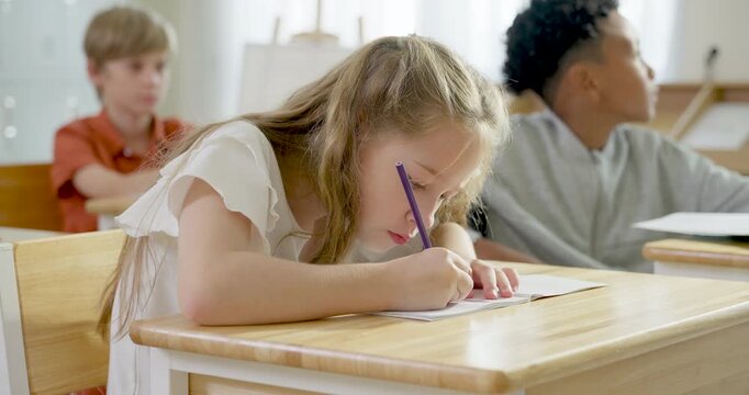 Blonde elementary girl writes in notebook during slow motion classroom exercise while multicultural classmates study behind, back to school activity builds confidence, attention, learning progress