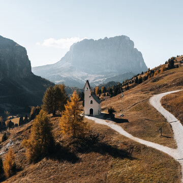 Aerial view of Cappella di San Maurizio surrounded by golden autumn trees and the majestic rocky peaks of the Dolomites in Selva di Val Gardena, Trentino-Alto Adige, Italy.