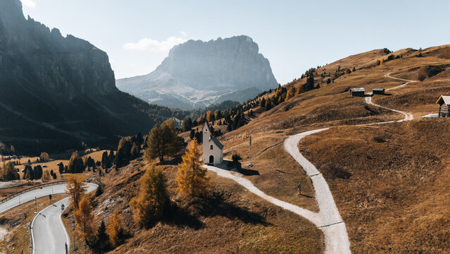 Aerial view of Cappella di San Maurizio nestled among golden autumn larches with the majestic Dolomites and winding mountain roads in Selva di Val Gardena, Trentino-Alto Adige, Italy.