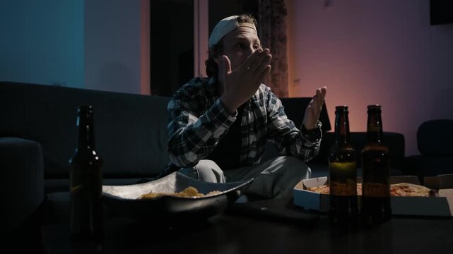 Medium wide shot of frustrated man emotionally reacting to sports defeat while watching television at night, disappointed fan gesture in cinematic apartment interior.