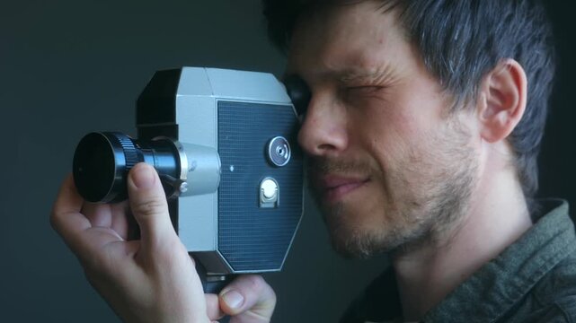 Filmmaker with a beard holding vintage camera, looking through viewfinder and adjusting lens to record nostalgic looking movie on dark background in studio. Cameraman shooting film with old camera