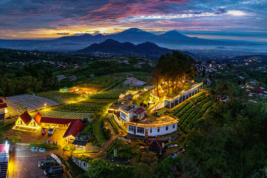 Aerial view of Silancur Highland with illuminated buildings and terraced fields against mountains under an orange sunset sky Magelang Regency, Central Java, Indonesia.