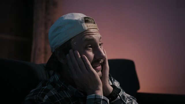 Medium close-up shot of disappointed man covering face after sports defeat while watching television at night, emotional frustration in cinematic apartment lighting.