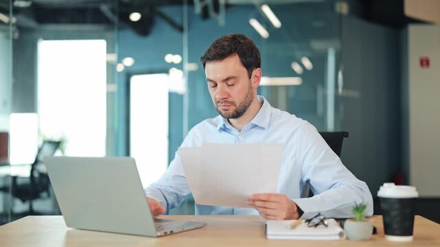 Concentrated male entrepreneur holds papers and reviews financial data while typing on laptop. Confident manager analyzing statistics at wooden workspace desk inside contemporary glass office.