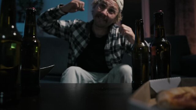 Foreground focus on beer bottles and pizza box while disappointed man sits blurred in background after sports defeat, drinking beer alone during emotional night television viewing.