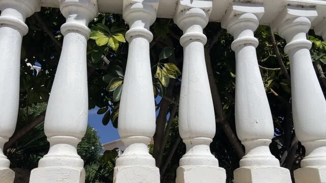 White classic balustrade on a sunny balcony, creating rhythmic architectural lines that frame leafy greenery and blue sky for a serene Mediterranean-style outdoor scene.