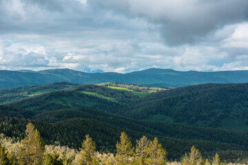 Scenic layered aerial top view from above to hilly woody vastness and dark silhouettes of mountains far away. Sunlight and shadows of clouds on forest hills under dramatic sky in changeable weather. © Daniil