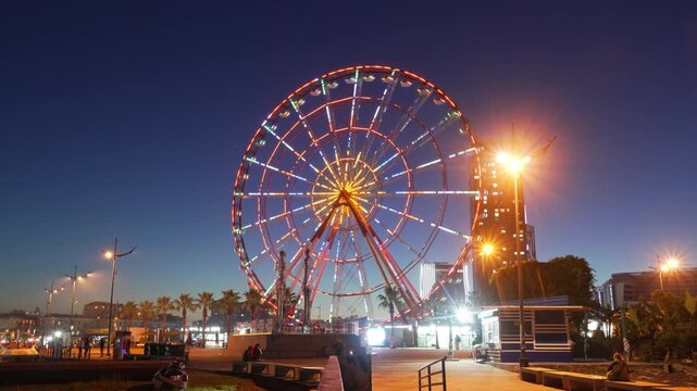 Panning timelapse of a spinning illuminated Ferris wheel on embankment in Batumi evening, sightseeing in Georgia, Adjara