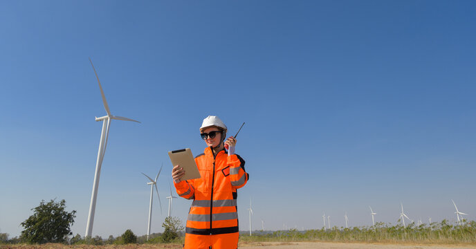 Female wind farm engineer reviewing data on digital tablet while using walkie talkie at wind energy site