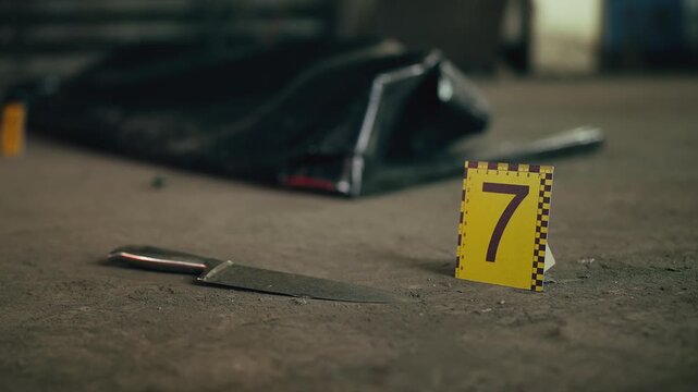 Close-up of knife and yellow evidence marker near body bag at crime scene