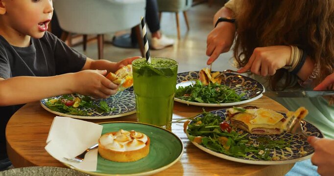 Plates with salad near toast and tart surround young siblings during family lunch. Boy eats burger piece while girl cuts salad with cheerful talk continuing