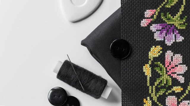 Black fabric, colorful floral cross-stitch embroidery, a spool of black thread and needle, black buttons, and white tailor's chalk on a clean white surface. Sewing. Close-up, flatlay.