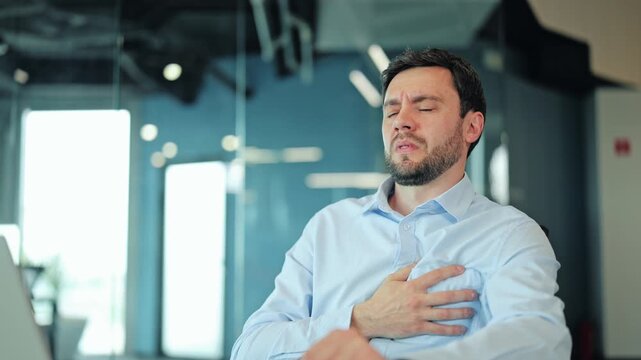 Overworked businessman sitting at desk with laptop and holding hand on chest, feeling sudden heart pain, illness, or panic attack symptoms. Sick corporate worker suffering from stress in office.