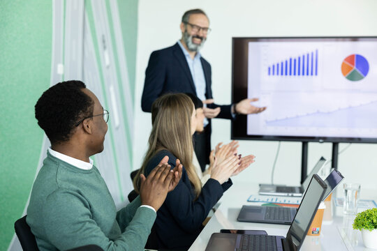 Business professionals applauding a male colleague presenting financial data and charts on a large screen during an office meeting