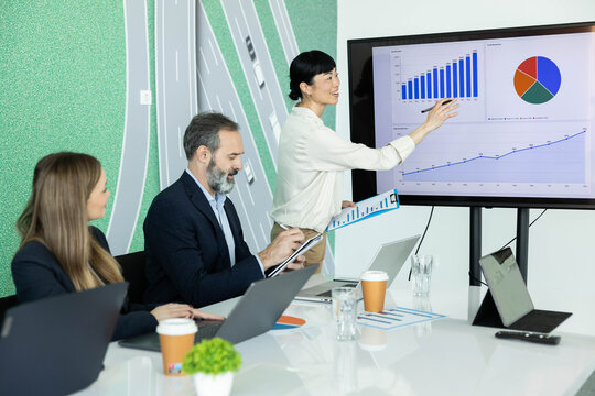 Asian woman is presenting growth charts on a large screen to diverse colleagues in a modern office boardroom