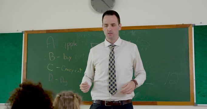 Male adult teacher gestures beside chalkboard during back to school first day, guiding elementary class through primary lesson notes while young student group listens with curiosity and readiness