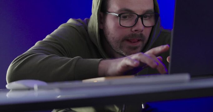 hooded man leaning toward a laptop and typing information on the keyboard in a blue neon environment. Suitable for cybersecurity, programming, data entry, online activity, IT work, and digital 