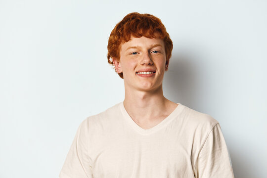 Young man with red hair and freckles wearing white t shirt smiling and looking at camera on plain white background. Casual portrait of confident happy male model.