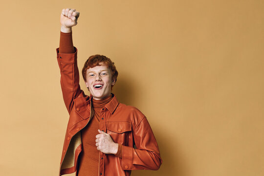Young man with red hair wearing orange jacket and turtleneck raising fist in celebration against plain beige background with smile and excitement.