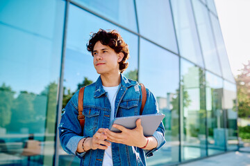 Young man wearing a denim jacket holding a digital tablet while standing outside a modern glass building. Looking forward © we.bond.creations