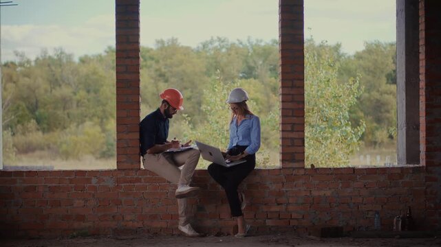 Engineers Sitting On Window Sill Of Unfinished Brick Building. Discussion Architecture Project And Scheme Documents On Construction Site