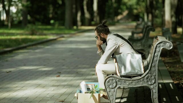 Fired Businessman Sitting On Bench Near Box With Personal Belongings. Dismissed Employee Thinking About Job Loss Crisis. Unemployment 