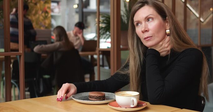 Dreaming woman sits in cafe near cappuccino and chocolate macaron on table. Female guest touches neck and tilts head looking at window with serious attention