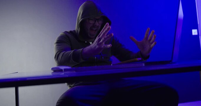hooded man screaming and holding his head in frustration while reacting to stressful laptop content. Suitable computer problems, cyber stress, hack failure, online frustration, burnout, technology