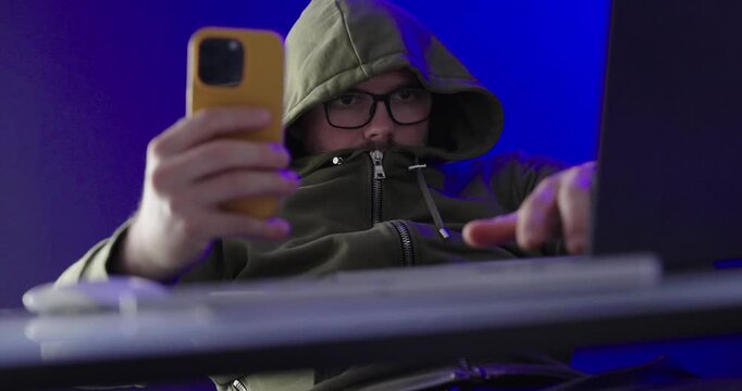 Hooded Man Hiding Face While Using Smartphone and Laptop