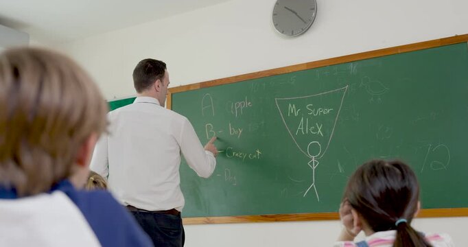 Male adult teacher writes math lesson on chalkboard while child raises yellow pencil to answer question, creating back to school elementary classroom discussion with confidence and active learning
