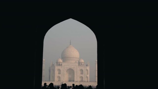 Majestic view of the Taj Mahal mausoleum in Agra, India, perfectly framed by the silhouette of an ancient archway as a crowd of tourists gathers at the famous historical landmark