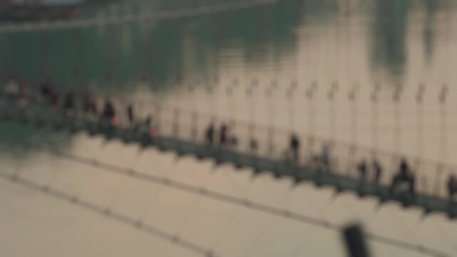 Blurry view of a crowd of unrecognizable people walking on a suspension bridge crossing a wide river, with the structure reflecting on the calm water surface during a tranquil day