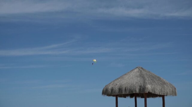 Thatched beach shelter beside open blue sky with distant parasail above tropical coastline and peaceful seaside atmosphere