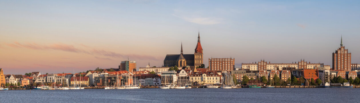 The Rostock skyline stretches along the Warnow River in warm evening light in Mecklenburg-Western Pomerania, Germany.