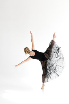 Full body rear view of a young female contemporary dancer in a black leotard and sheer black polka dot tulle skirt performing a dramatic arabesque-style high kick on one leg